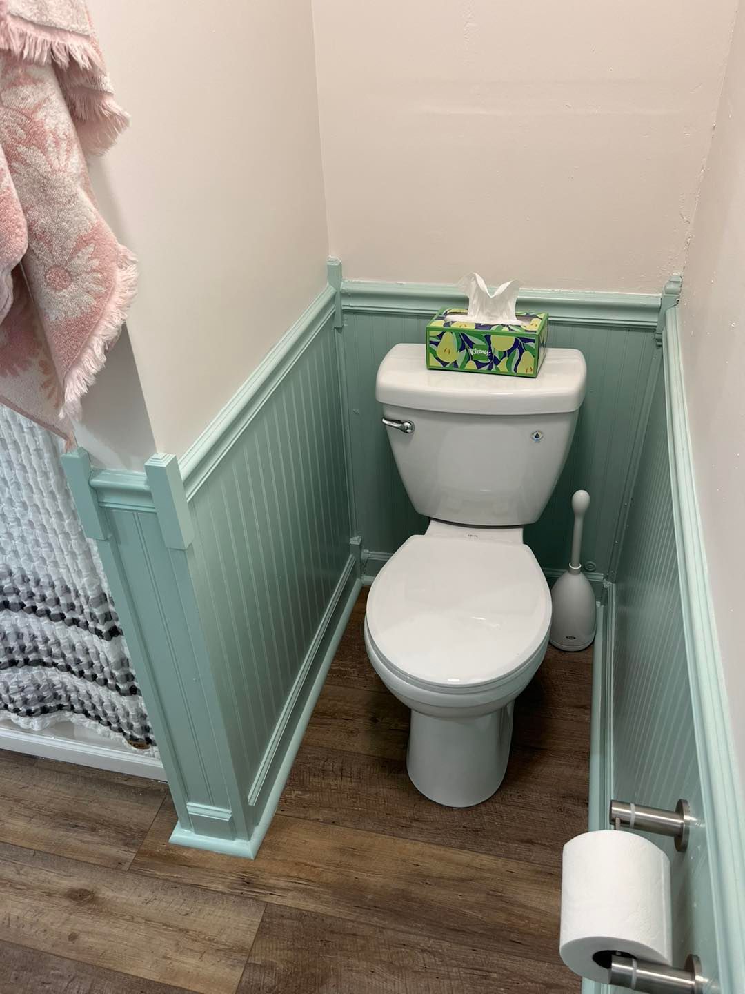 Small bathroom with a white toilet, teal wainscoting, and wood-look flooring; tissue box on tank.
