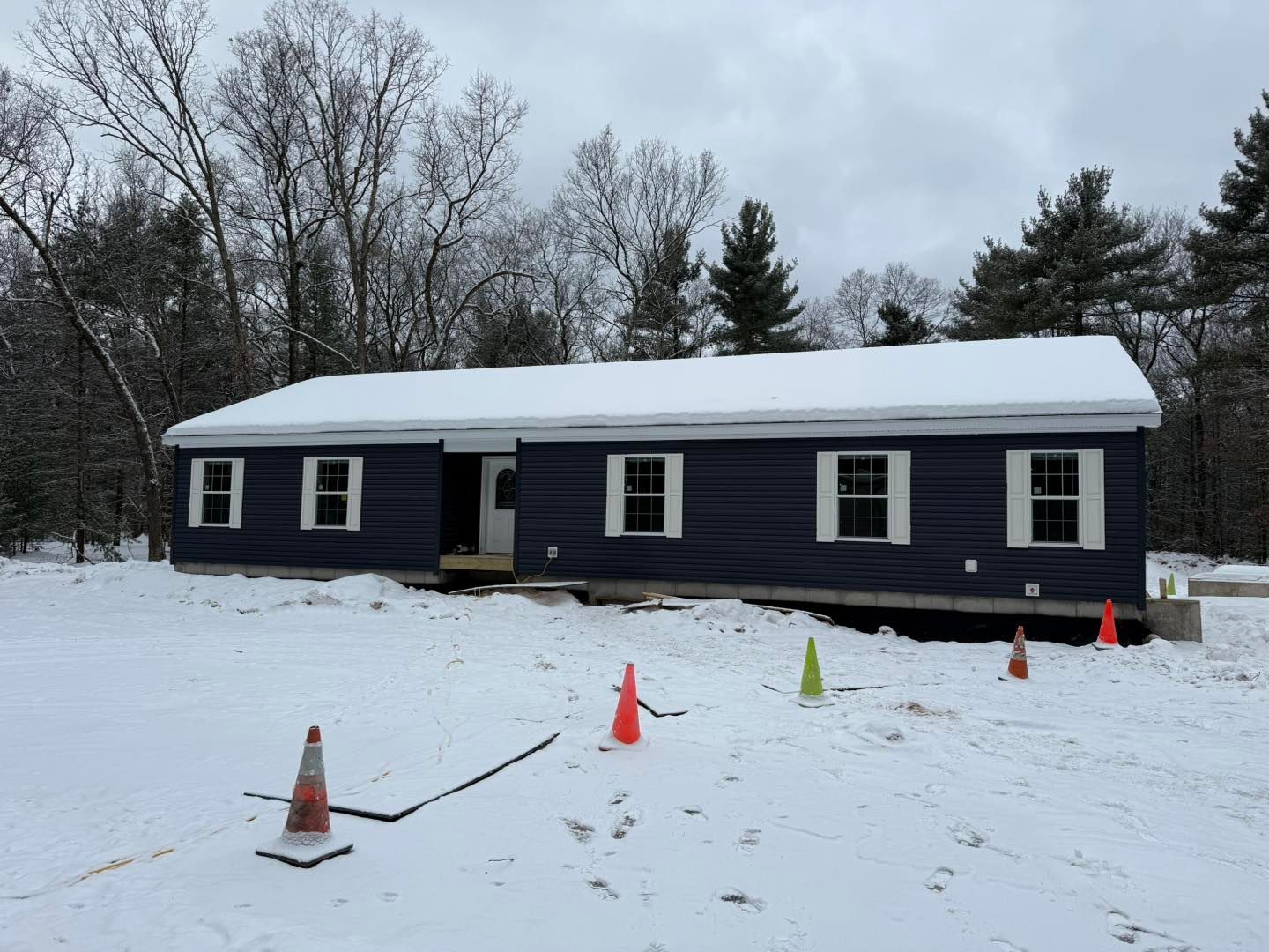 Dark blue single-story house in snowy yard, with cones and a snow-covered roof, surrounded by bare trees.