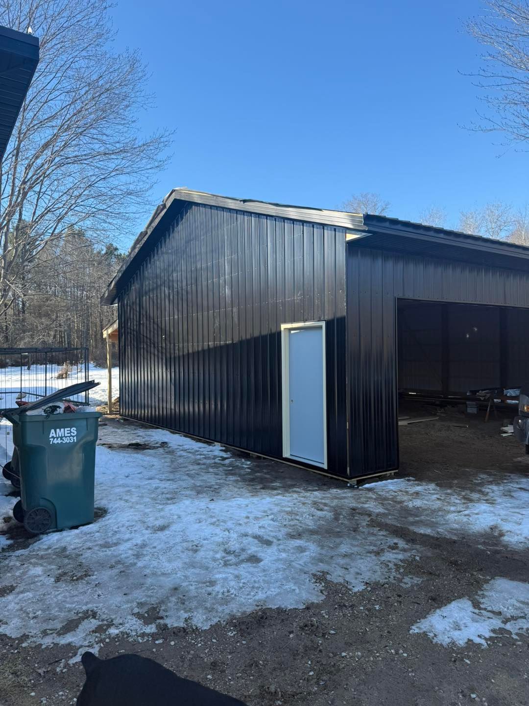 Black shed with open garage door on a snowy driveway, with a green trash bin nearby.