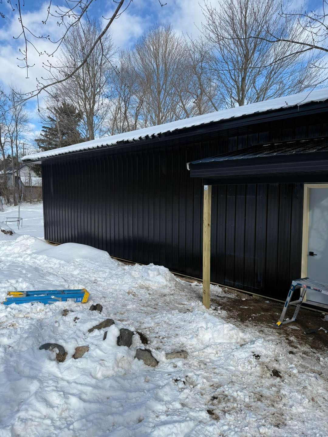 Black shed with snow-covered roof in a snowy yard, trees and blue sky behind it.