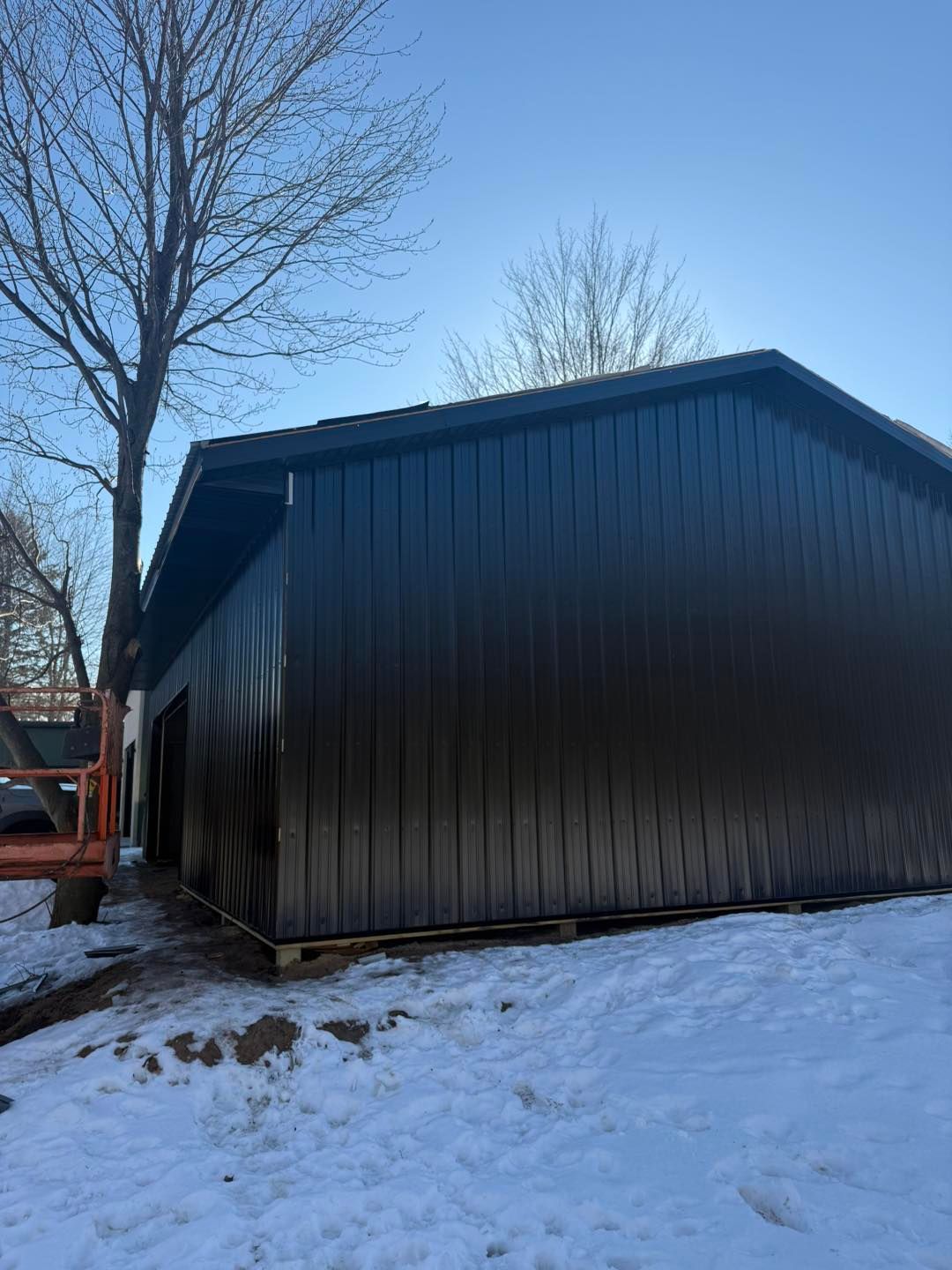 Black metal shed beside a snowy yard with bare trees and an orange vehicle in the background
