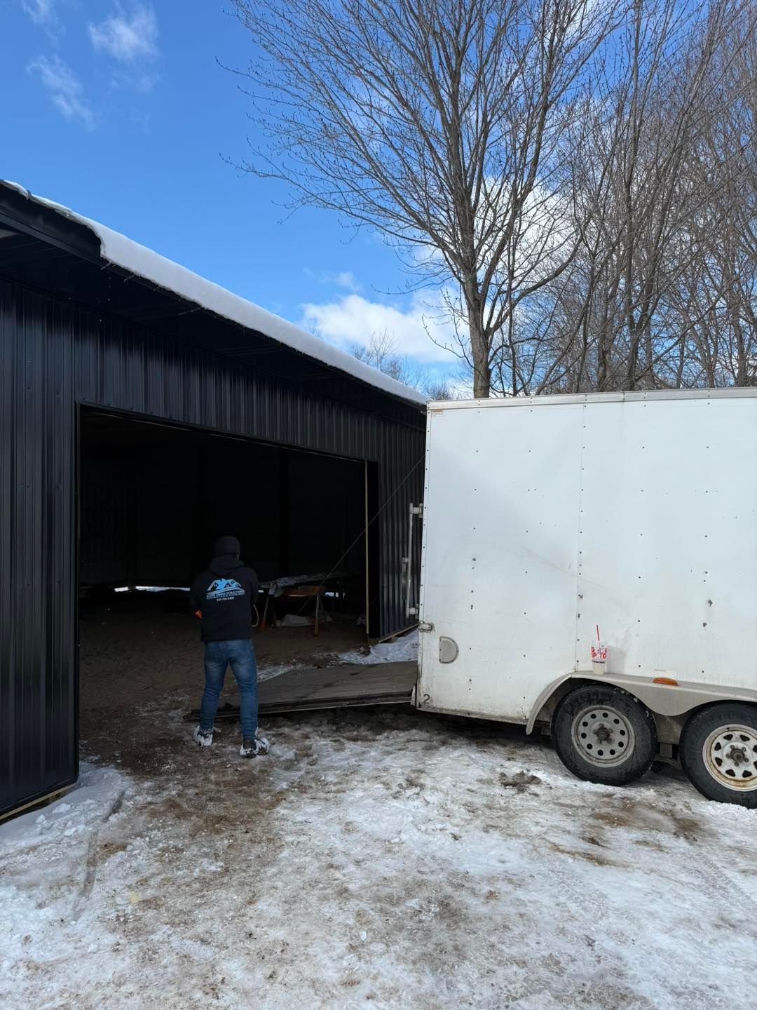 Person in a black jacket stands by a barn entrance beside a white trailer on a snowy day.