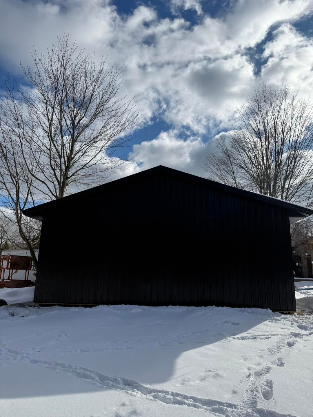 Snowy yard with a dark barn-like building beneath bare trees and a cloudy sky