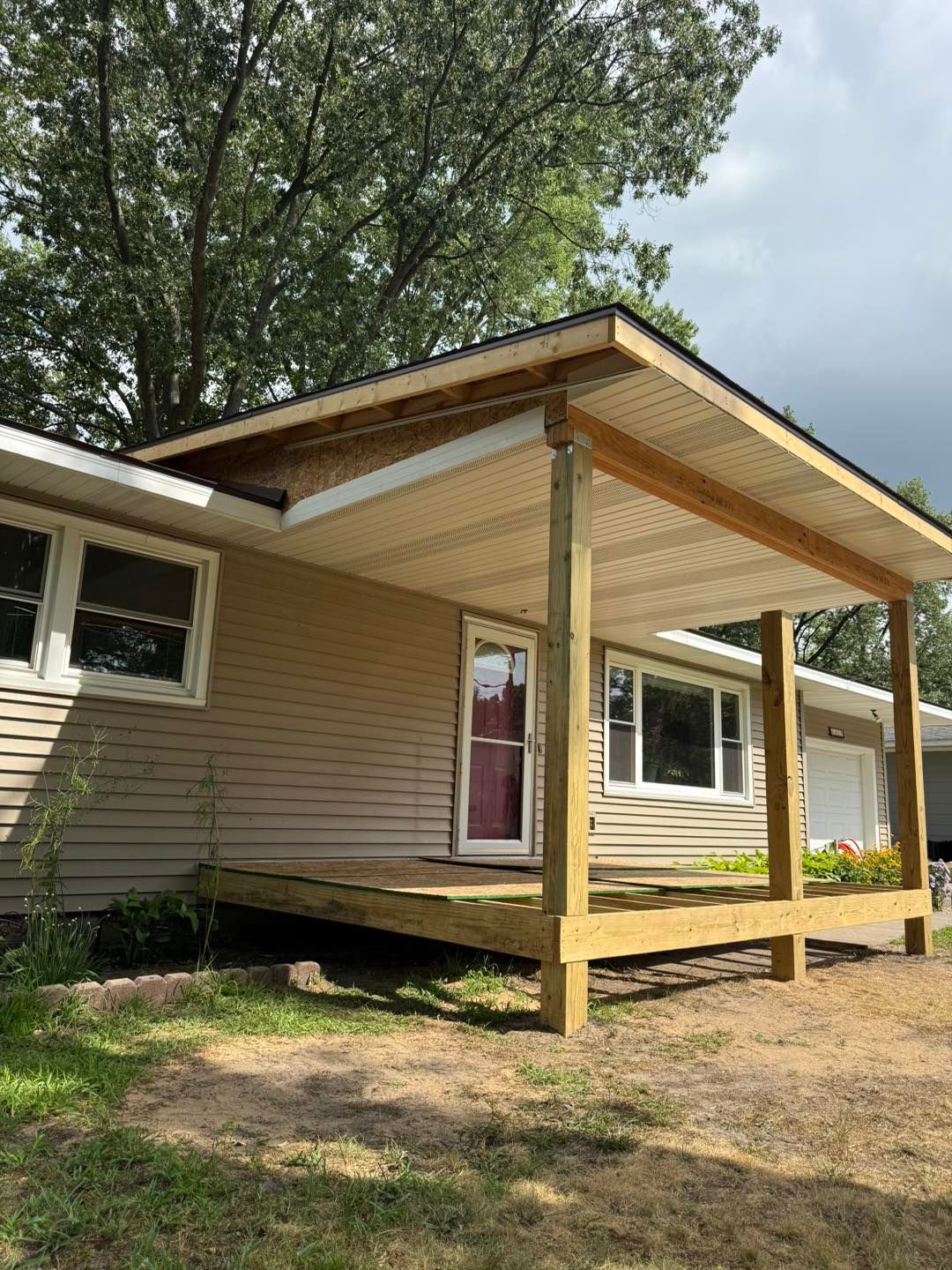 Small beige house with a covered front porch and wooden support posts under tall trees.