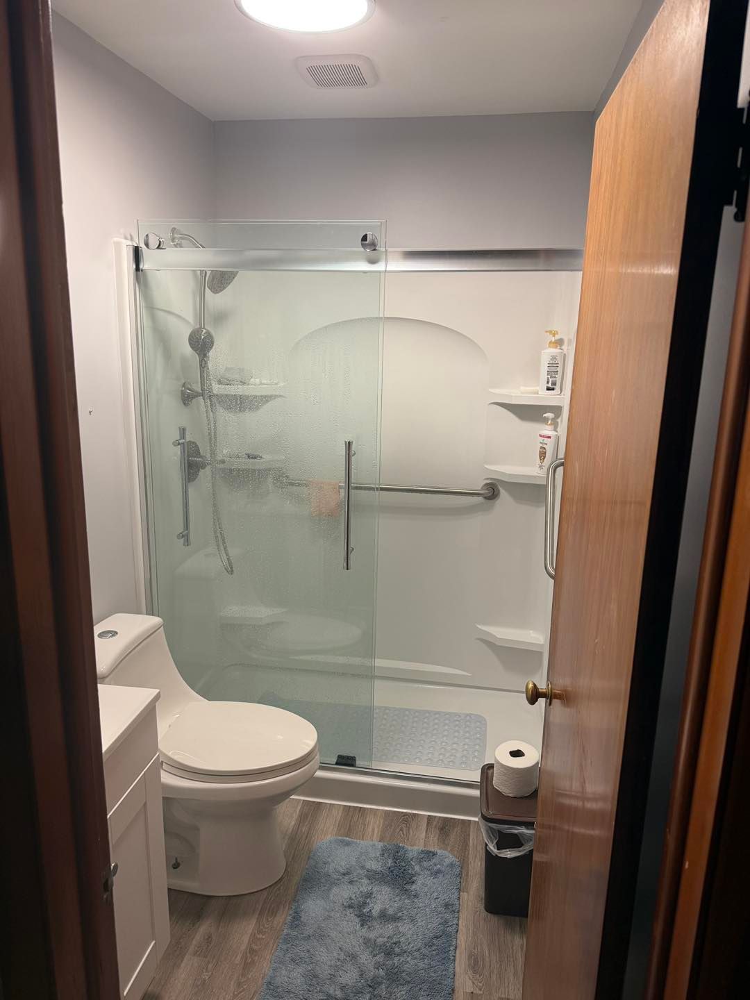Accessible bathroom with toilet, blue bath mat, and glass walk-in shower with grab bars.