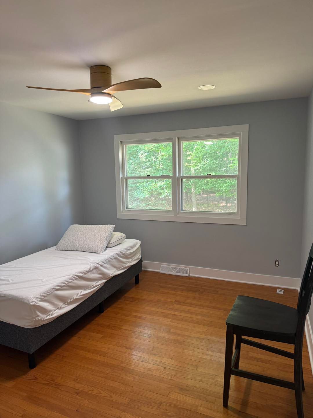 Small bedroom with a bed, chair, ceiling fan, and two windows overlooking greenery
