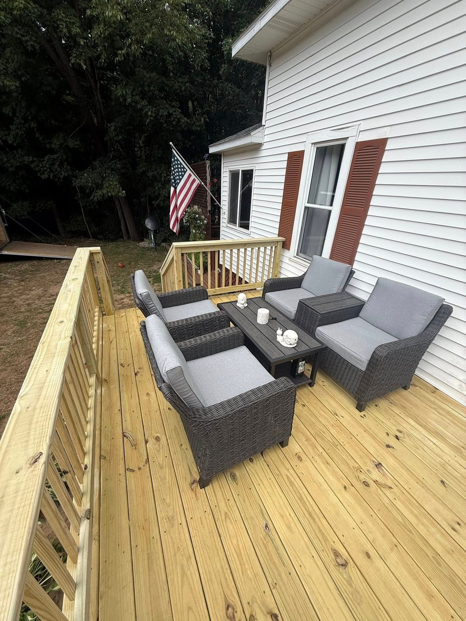 Deck with wicker patio chairs and a table beside a white house, with an American flag and wooded yard.