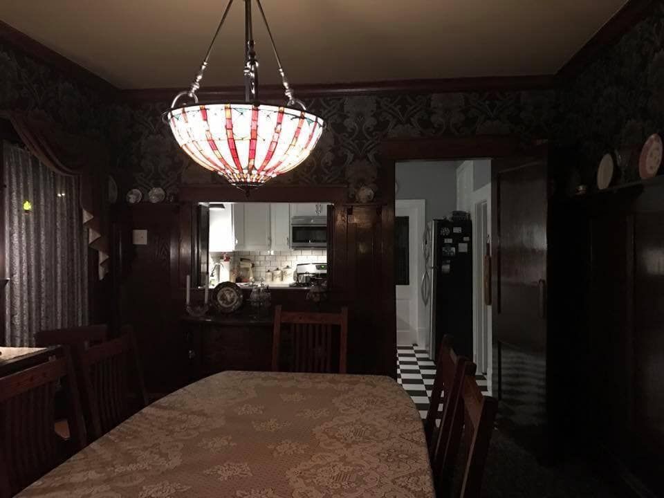 Dim dining room with a table, hanging red-and-white light, and a mirror reflecting a kitchen.