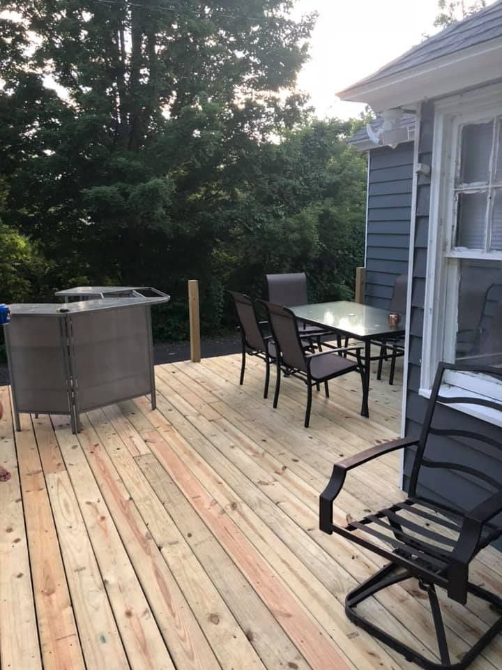 Wooden backyard deck with patio table, chairs, and a grill beside a house and trees.
