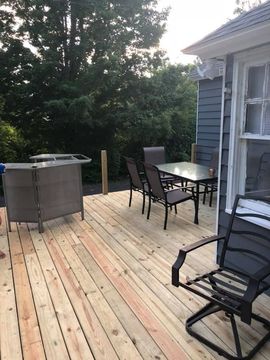 Wooden backyard deck with patio table, chairs, and a grill beside a house and trees.