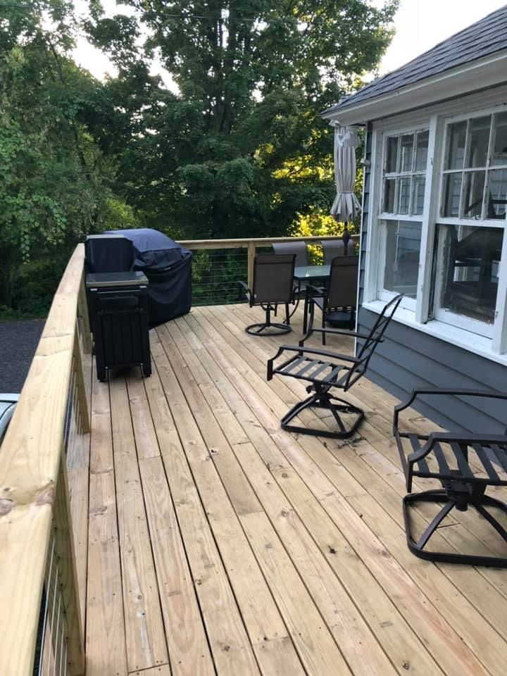 Wooden deck with grill, patio chairs, and glass doors beside a house, bordered by trees