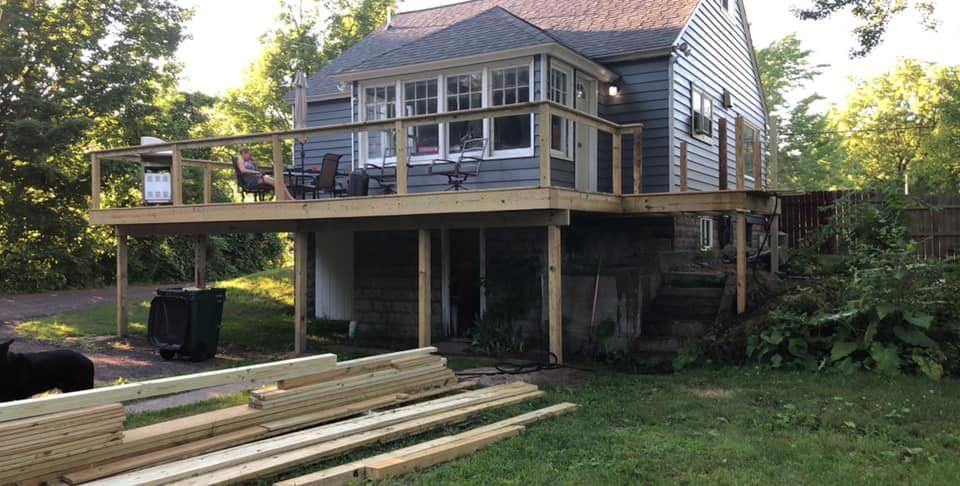 Raised house with a wraparound deck and stairs, viewed from a grassy yard with lumber in the foreground