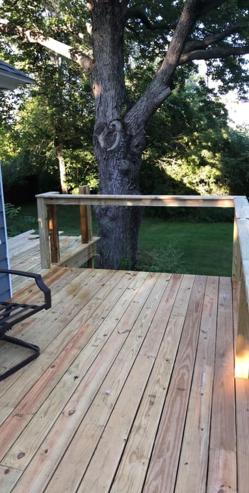 Wooden deck with railings around a large tree in a grassy backyard at dusk.