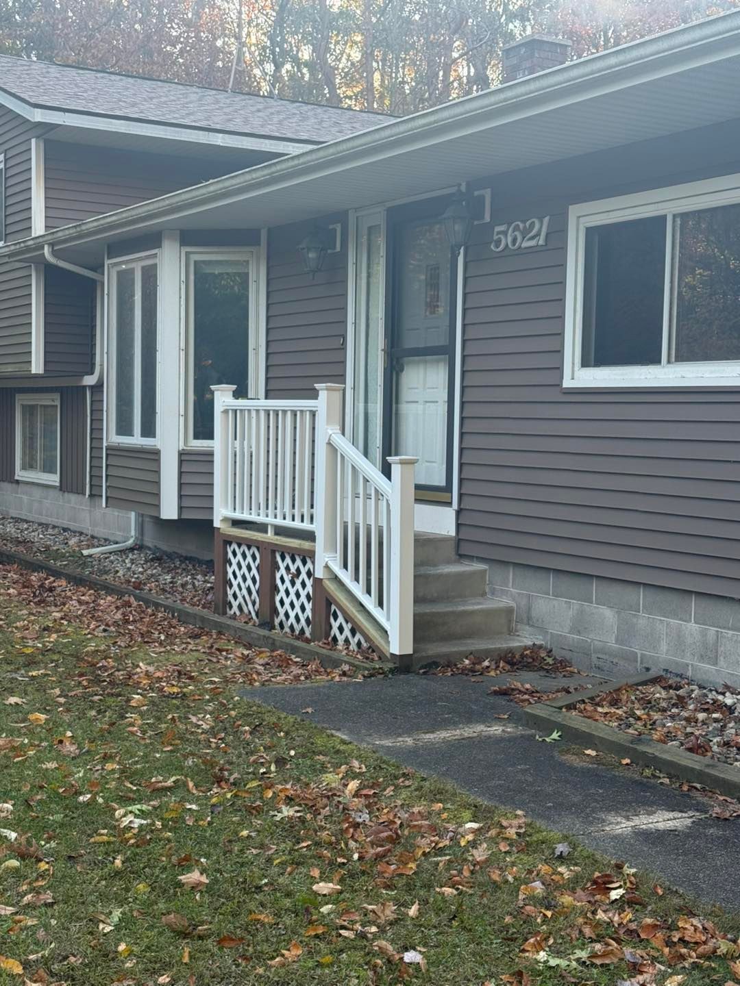 Gray house entry with small white porch, concrete ramp, and autumn leaves on the lawn