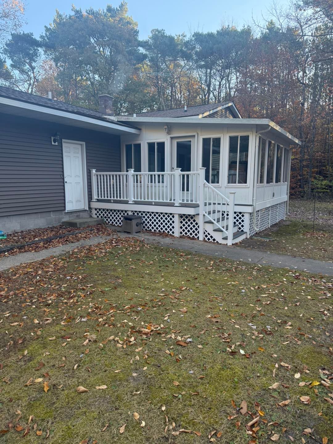 White porch on gray house with lattice skirting, set in a grassy yard with autumn trees in the background