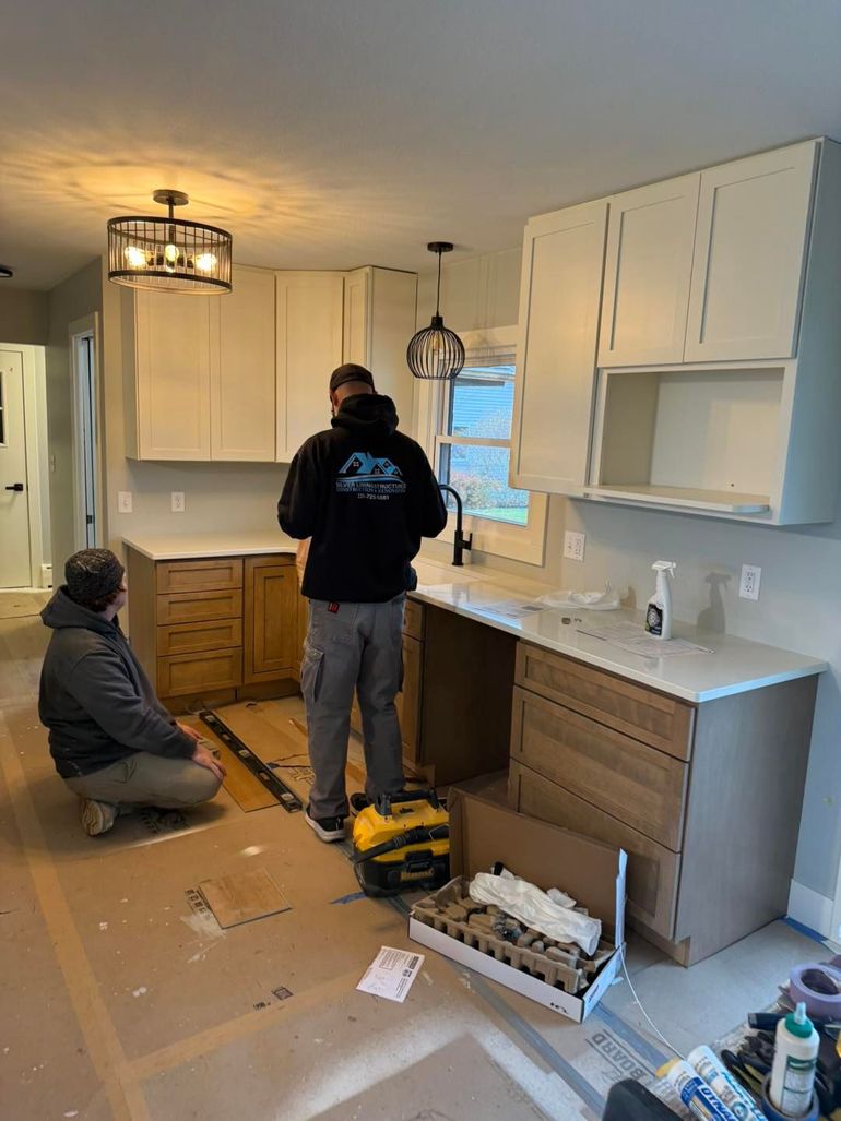 Two workers installing kitchen cabinets and countertops in a bright, unfinished kitchen.