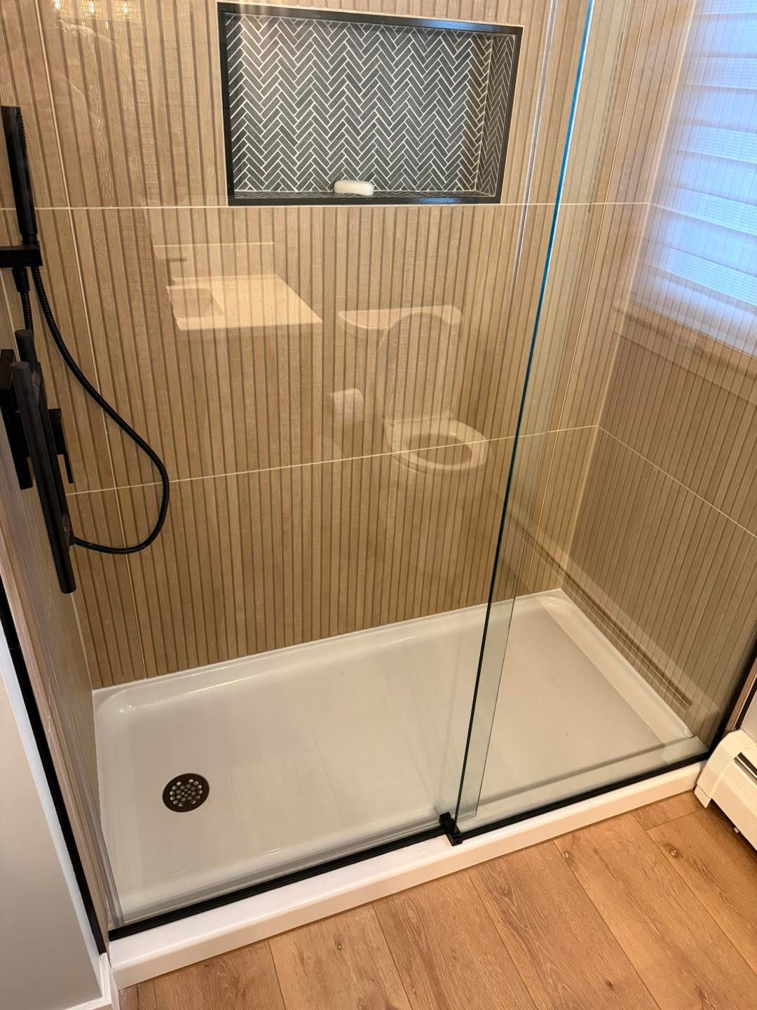 Glass-enclosed shower with white tile base, beige walls, and wood floor outside