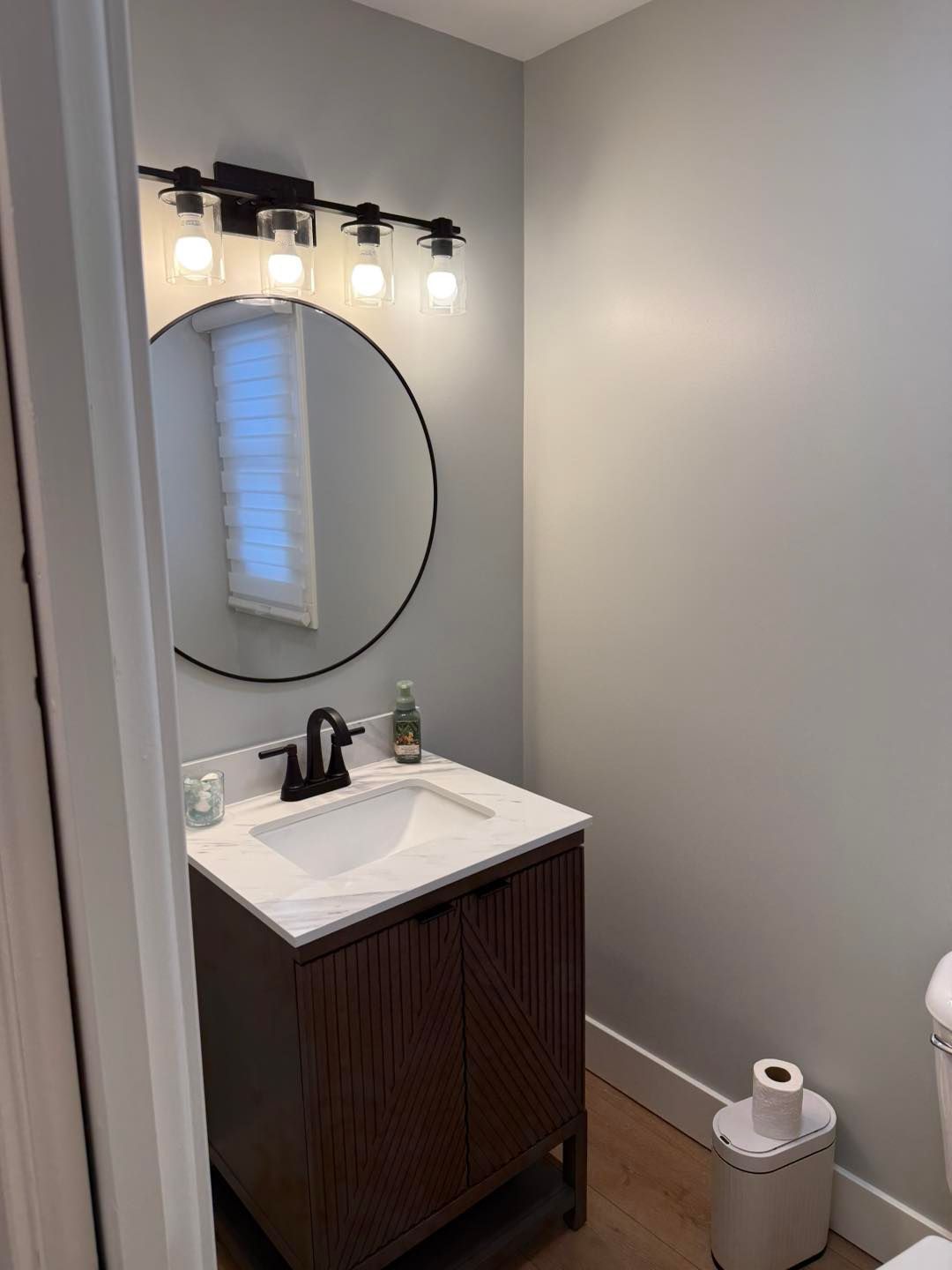 Small bathroom vanity with round mirror, wall lights, white sink, and toilet paper holder.