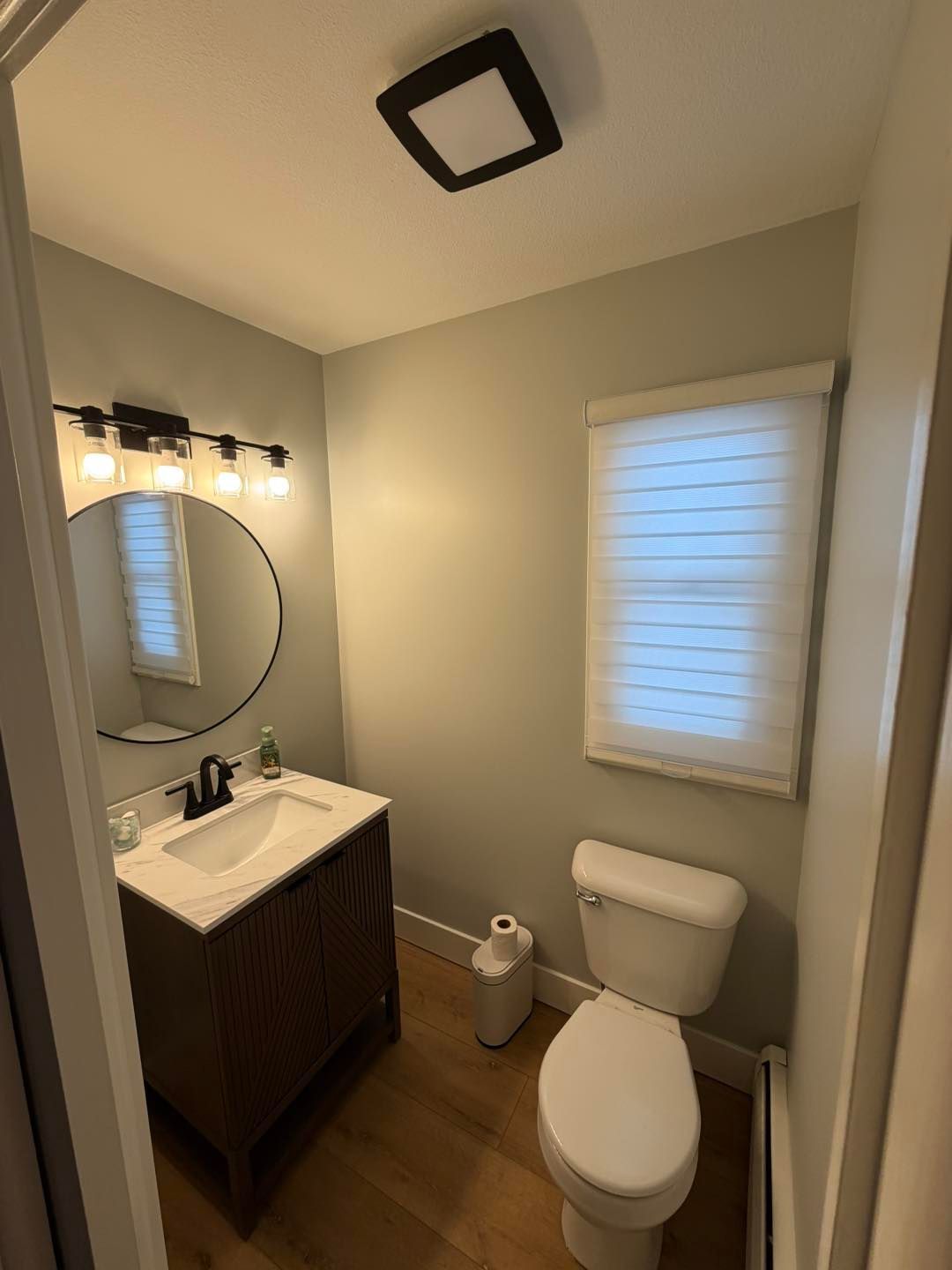 Small bathroom with vanity, round mirror, toilet, window, and wood flooring.