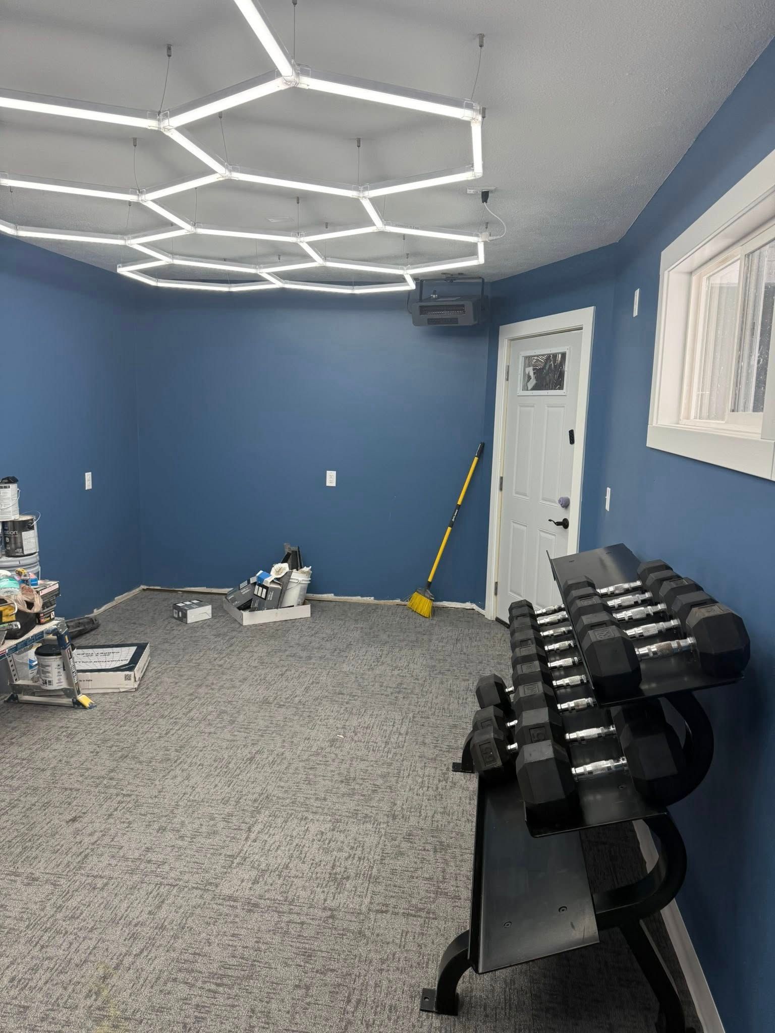 Blue home gym with hex ceiling lights, dumbbells, exercise equipment, and a white door.