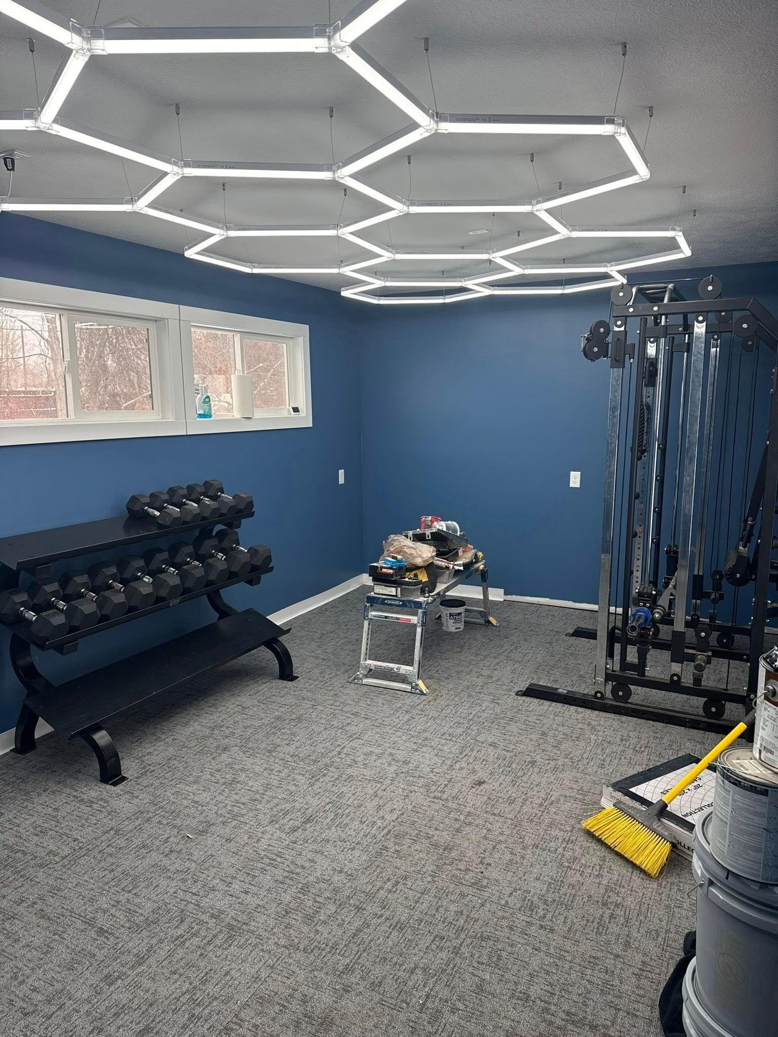 Small gym room with dumbbells, cable machine, bench, and blue walls under hexagonal ceiling lights.