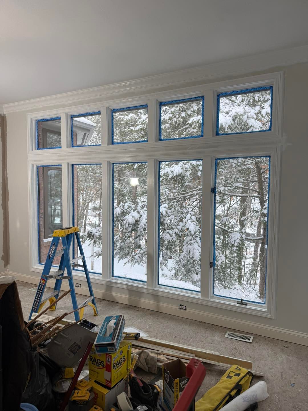 Snowy view through large windows in a room under renovation, with a blue ladder and tools in the foreground