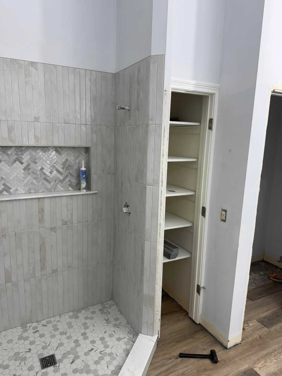 Tiled bathroom shower with built-in shelf and an open white linen cabinet beside it.