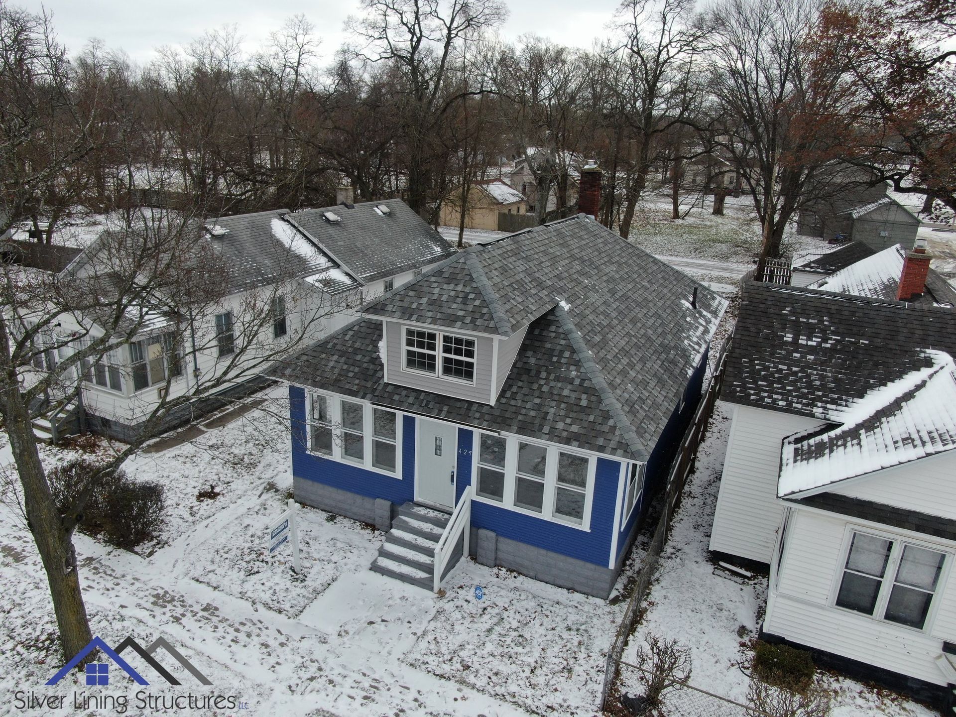 Snow-covered blue house in a residential neighborhood with bare trees and neighboring homes.