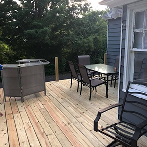 Wooden deck with black patio chairs and table beside a house, with a grill on the left.