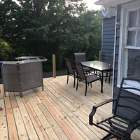 Wooden deck with black patio chairs and table beside a house, with a grill on the left.