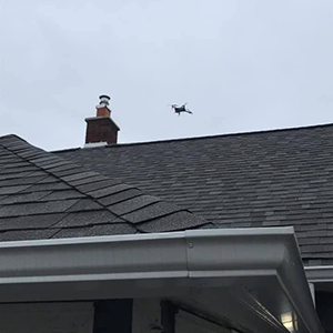 Small drone flying above a gray shingled roof and chimney under an overcast sky