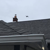 Small drone flying above a gray shingled roof and chimney under an overcast sky