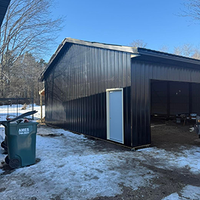 Black metal shed with open garage bay in snowy yard, with a green trash bin in front.