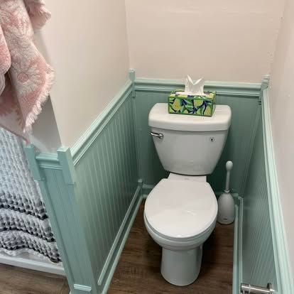 Small bathroom with a toilet, teal wainscoting, wood-look floor, and a toilet paper roll on the right.