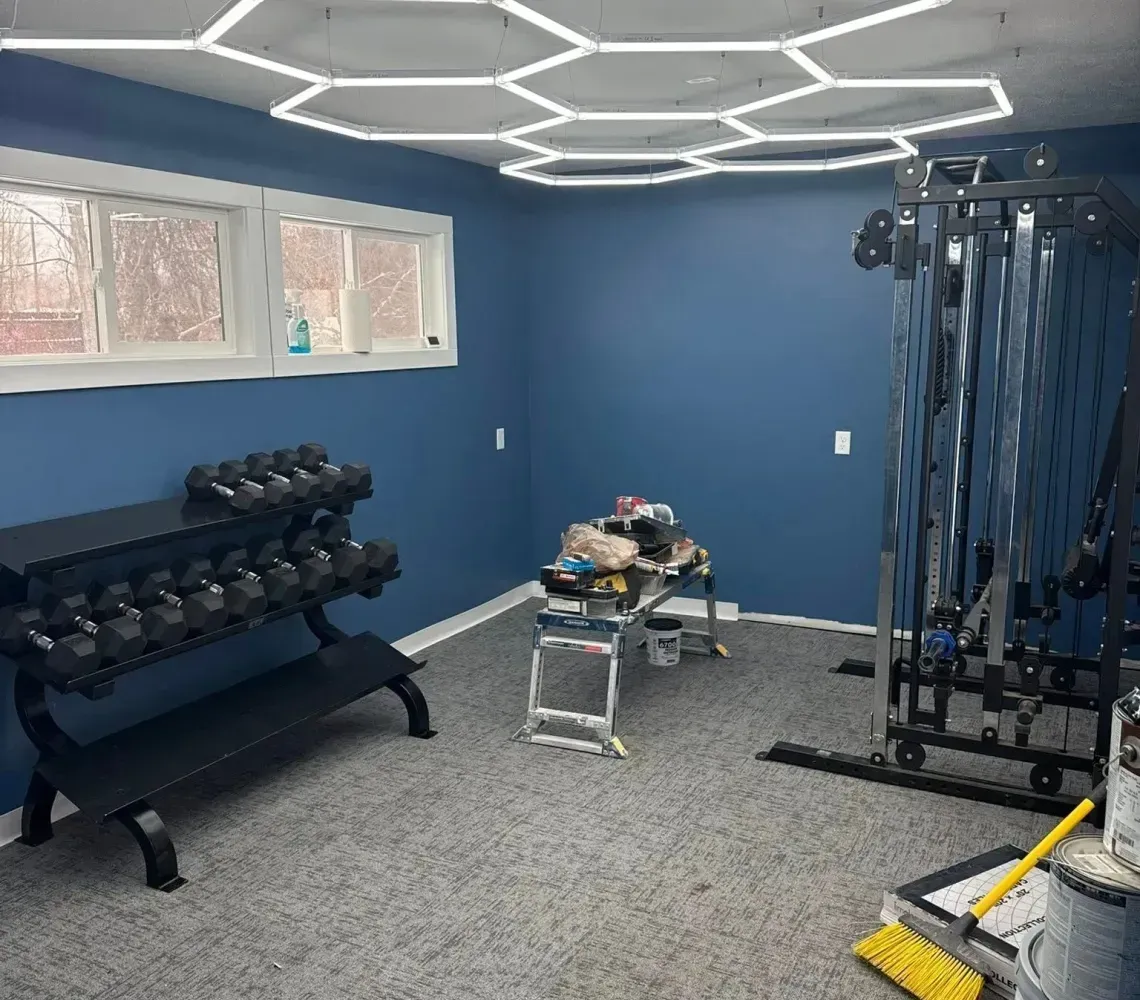 Small gym room with dumbbells, cable machine, bench, and blue walls under hexagonal ceiling lights.