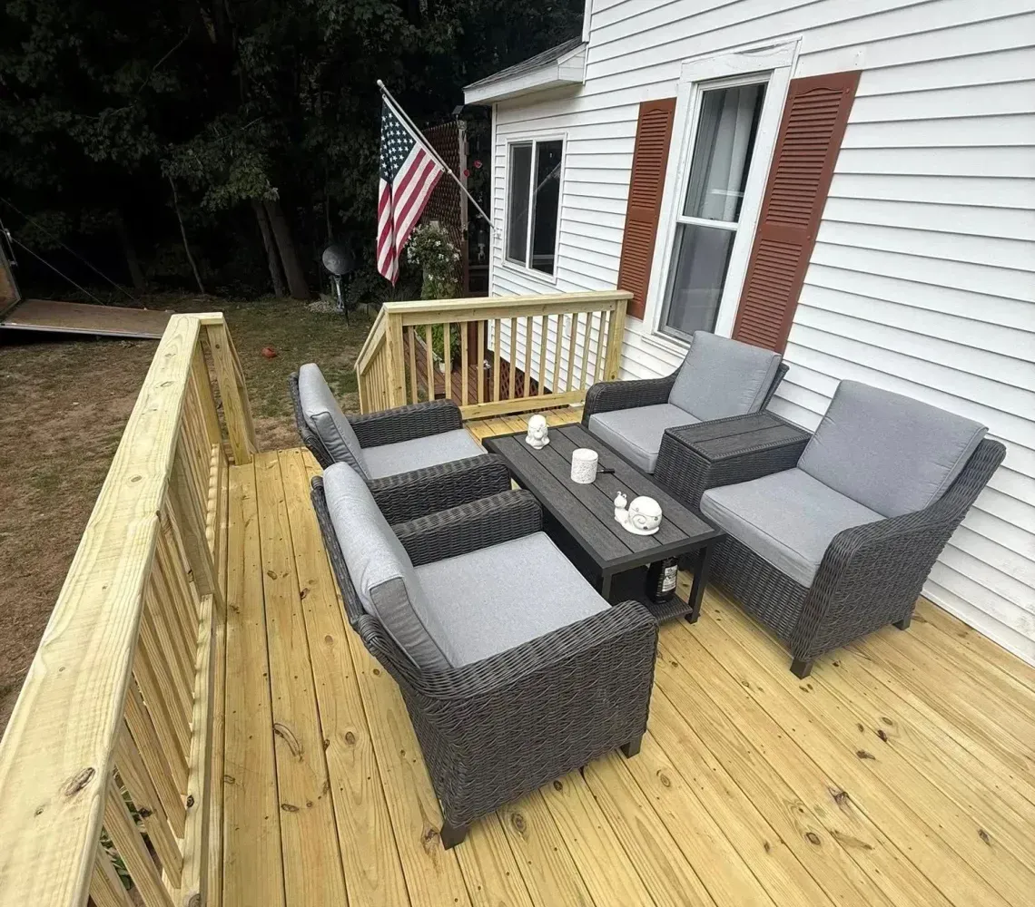 Wooden deck with gray wicker patio furniture, railing, and an American flag beside a white house