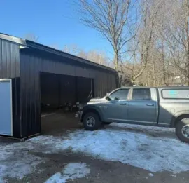 Gray pickup truck parked beside an open dark metal garage on a snowy driveway under a clear blue sky