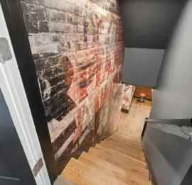 Narrow stairwell with wooden steps, gray walls, and a brick mural leading downward to a small room