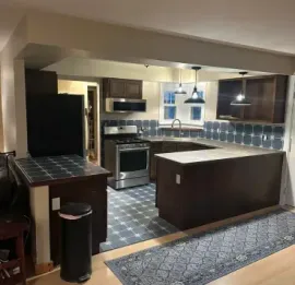 Modern kitchen with dark cabinets, central island, tiled backsplash, and gray rug leading in from a living area