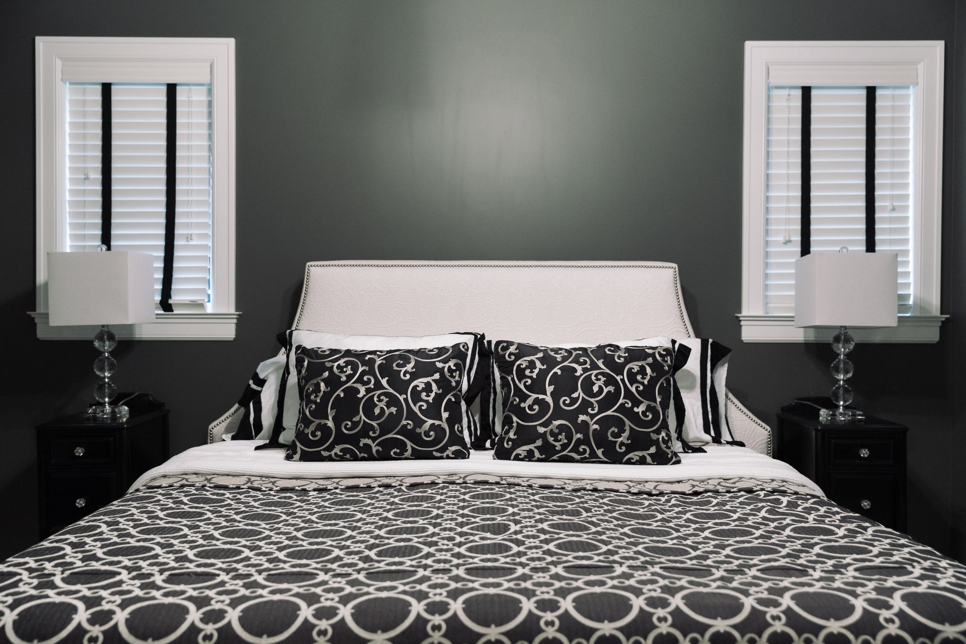 Dark bedroom with bed, two nightstands, two lamps, and two windows.