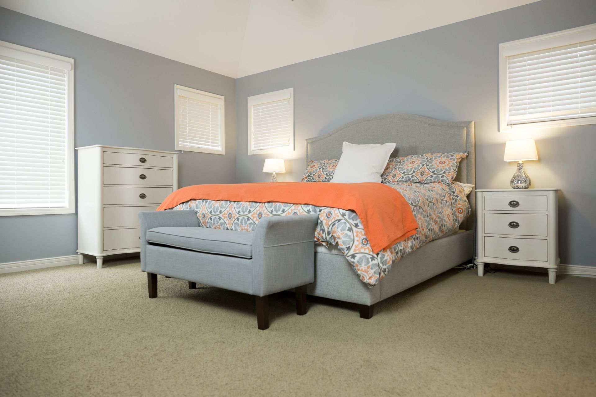 Bedroom with gray walls, bed, dresser, and bench. Orange blanket on bed, two windows.