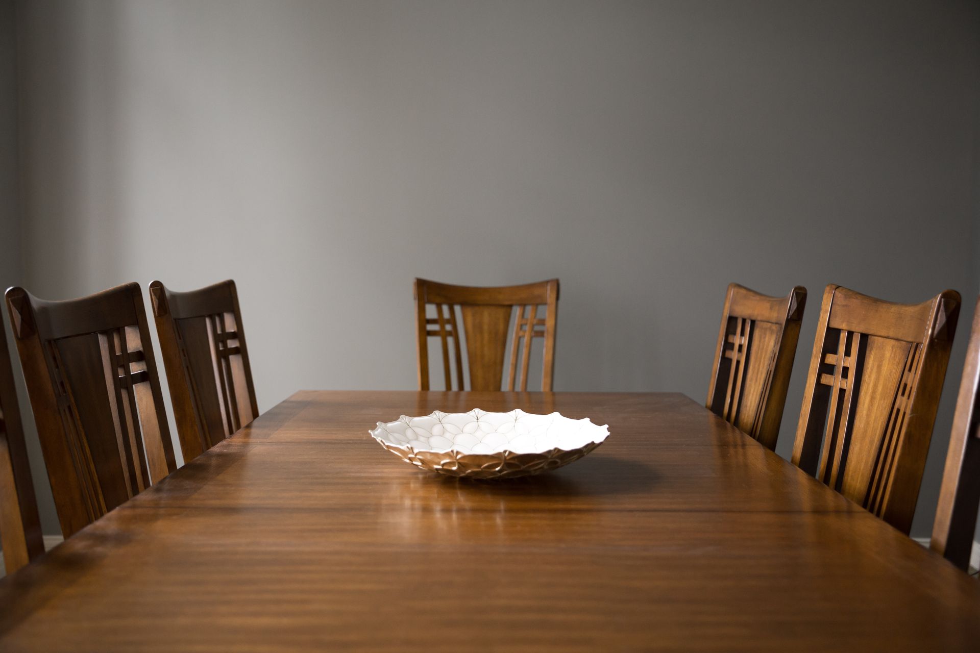 Wooden dining table with six chairs, a decorative bowl in the center. Neutral gray wall.
