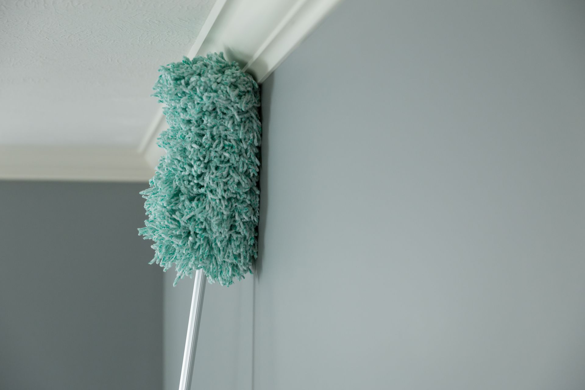 A teal duster cleaning a white crown molding on a gray wall.