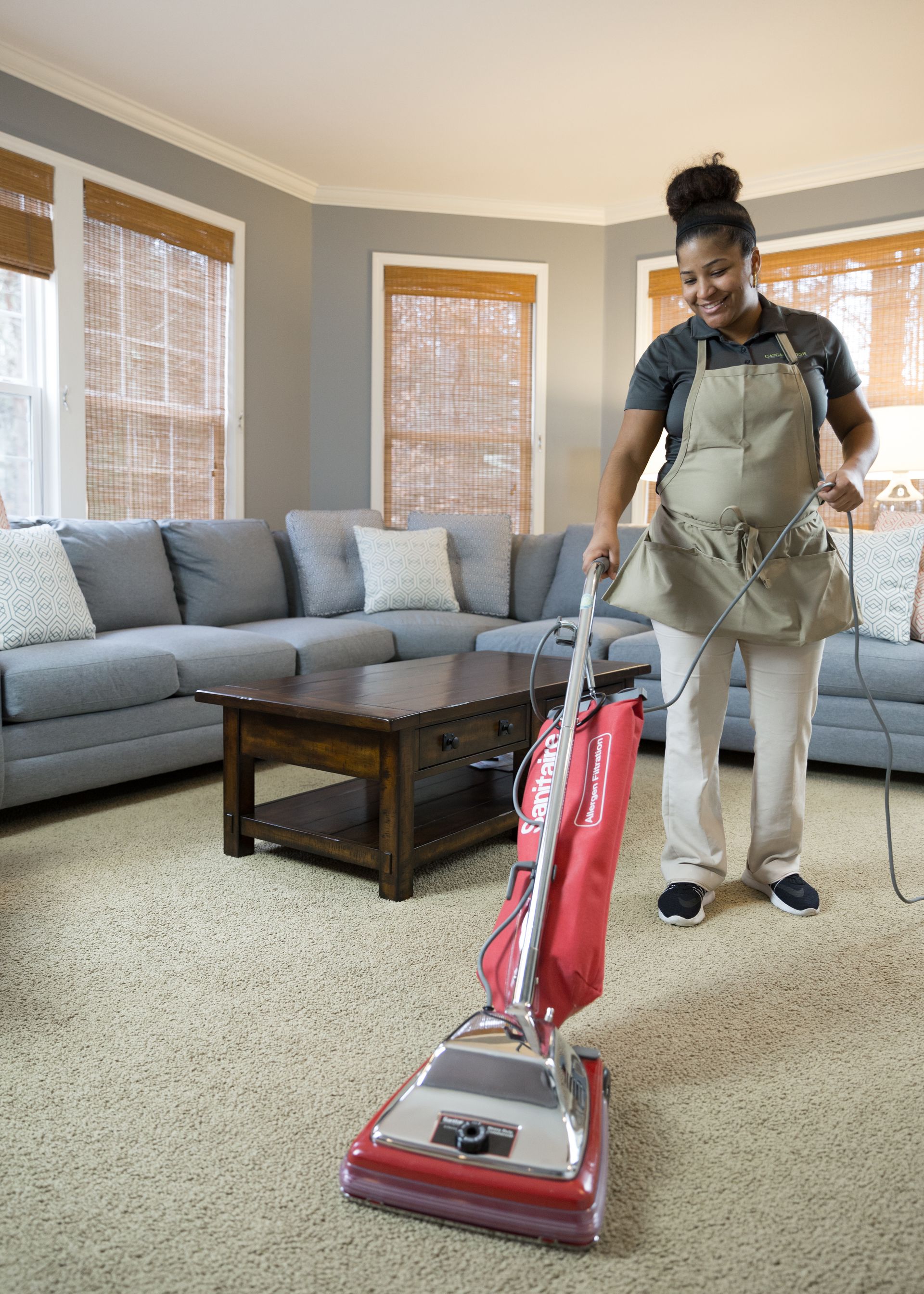 Woman vacuuming carpet in a living room, wearing apron and smiling.