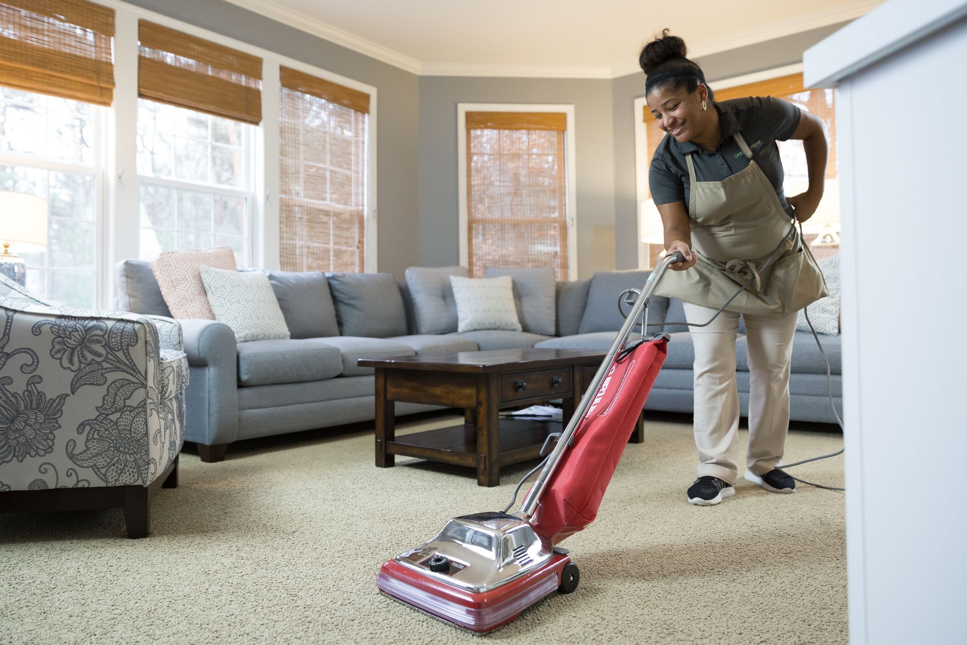 Woman vacuuming a carpet in a living room, wearing an apron, smiling.