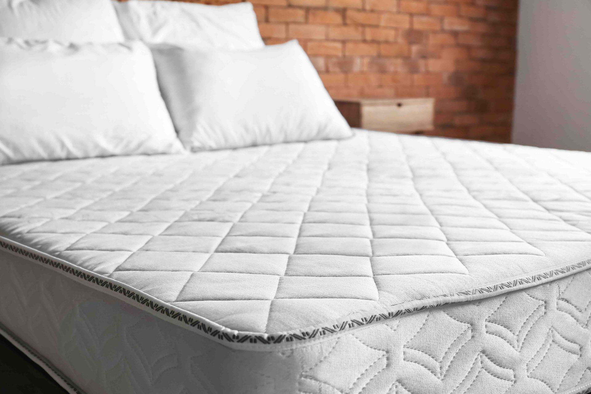 White quilted mattress with white pillows on a bed, against a brick wall.