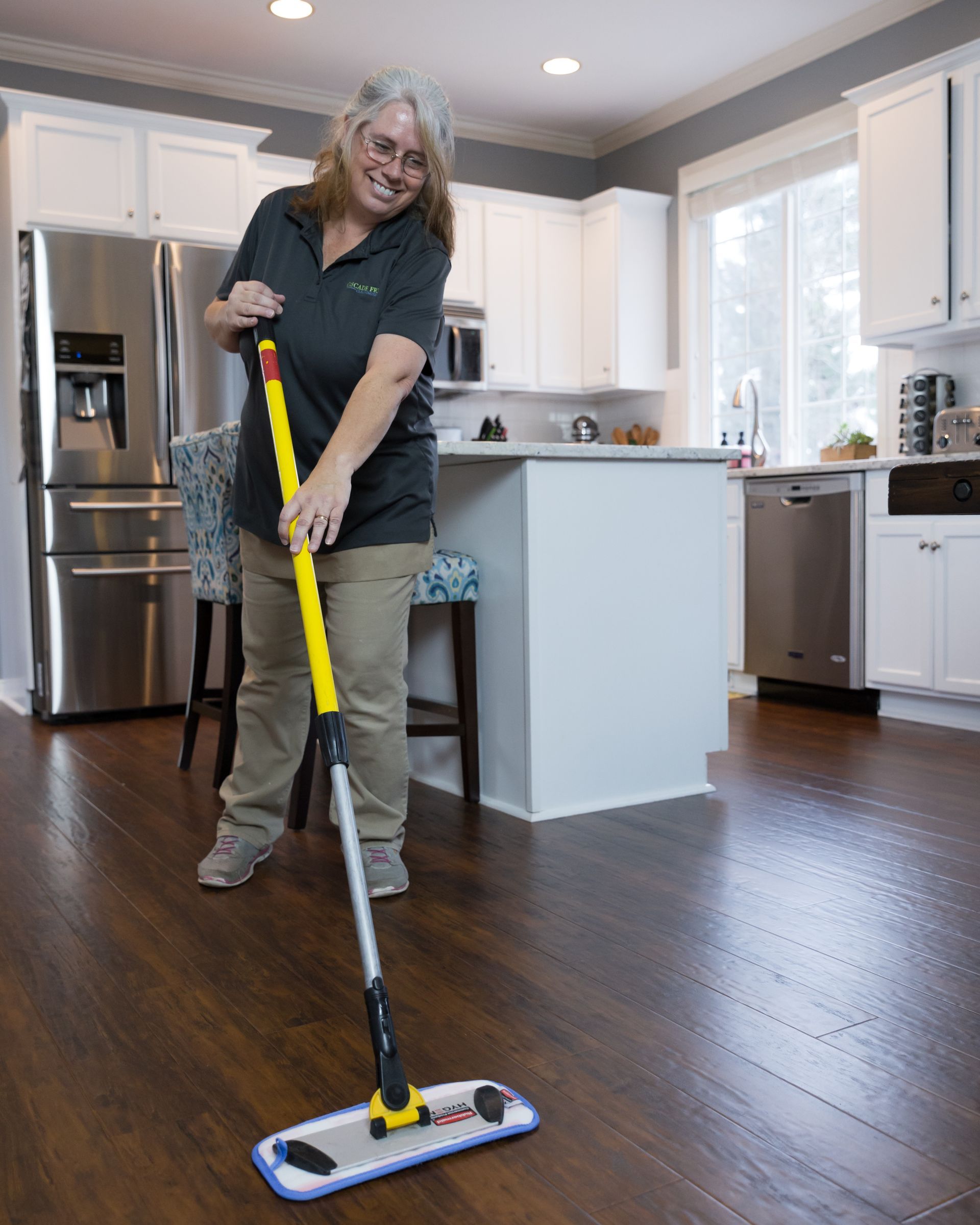 Woman mopping a wood floor in a kitchen, smiling. She is holding a yellow and silver mop.