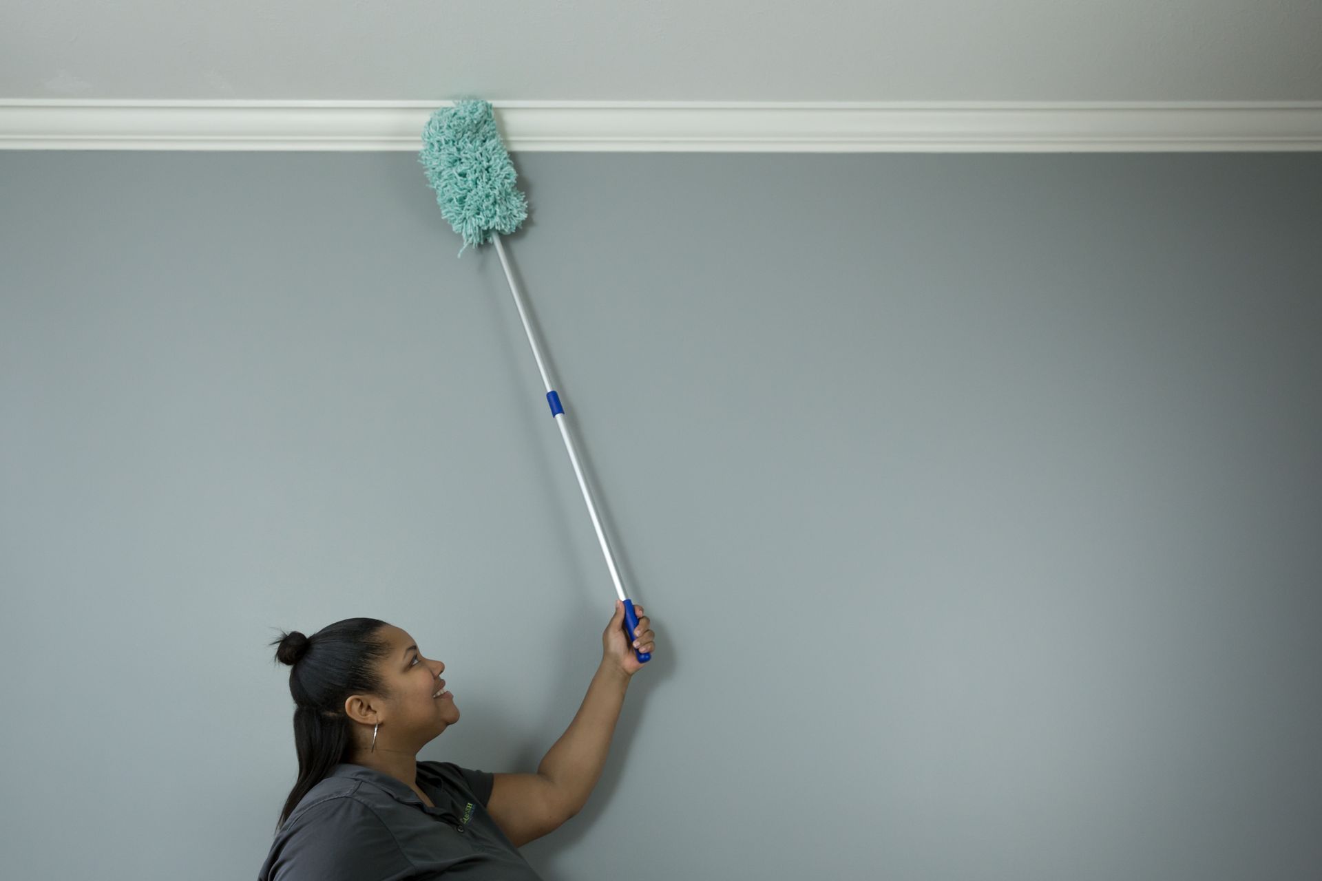 Woman dusting crown molding with an extendable duster. Grey wall and molding.
