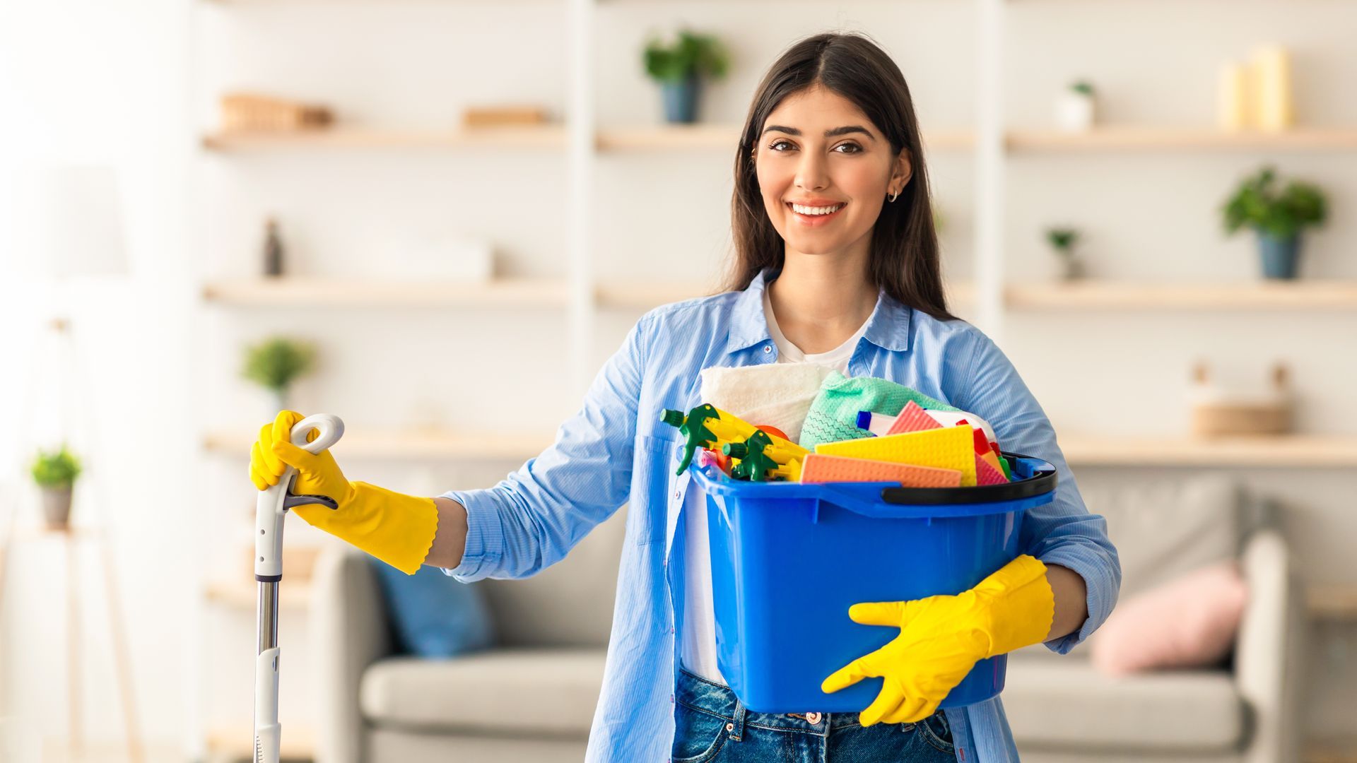 Woman in yellow gloves smiles, holding a bucket of cleaning supplies and a mop in a living room.