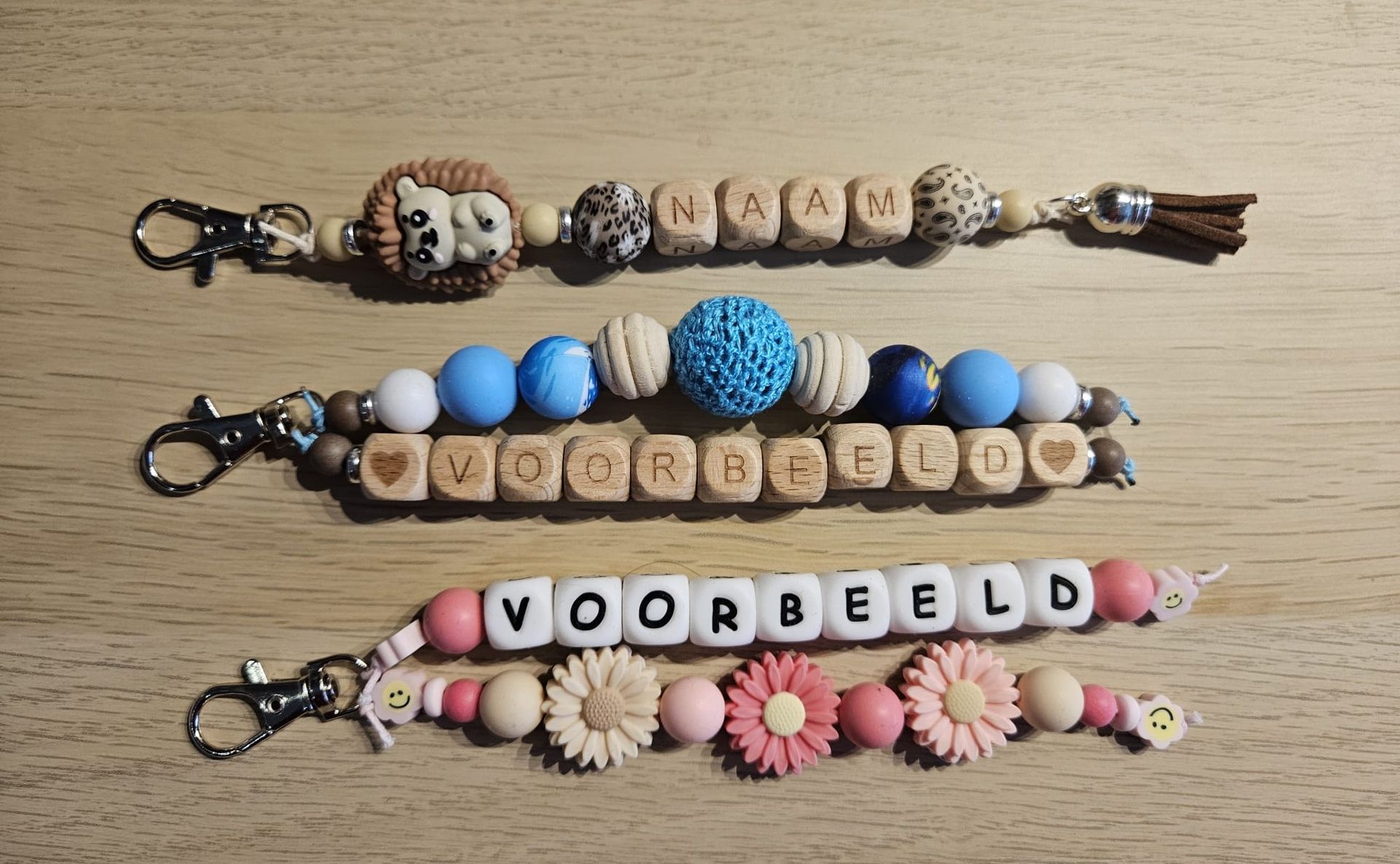Drie kralenarmbanden met kwastjes, elk met een woord erop, gerangschikt op een beige achtergrond.
